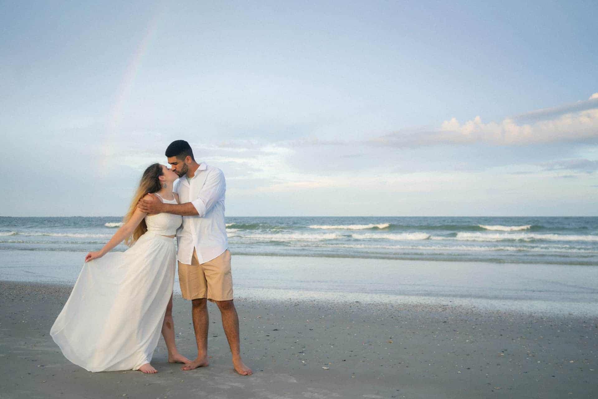 Couple sharing a kiss on the beach during sunset, wedding attire, ocean background.