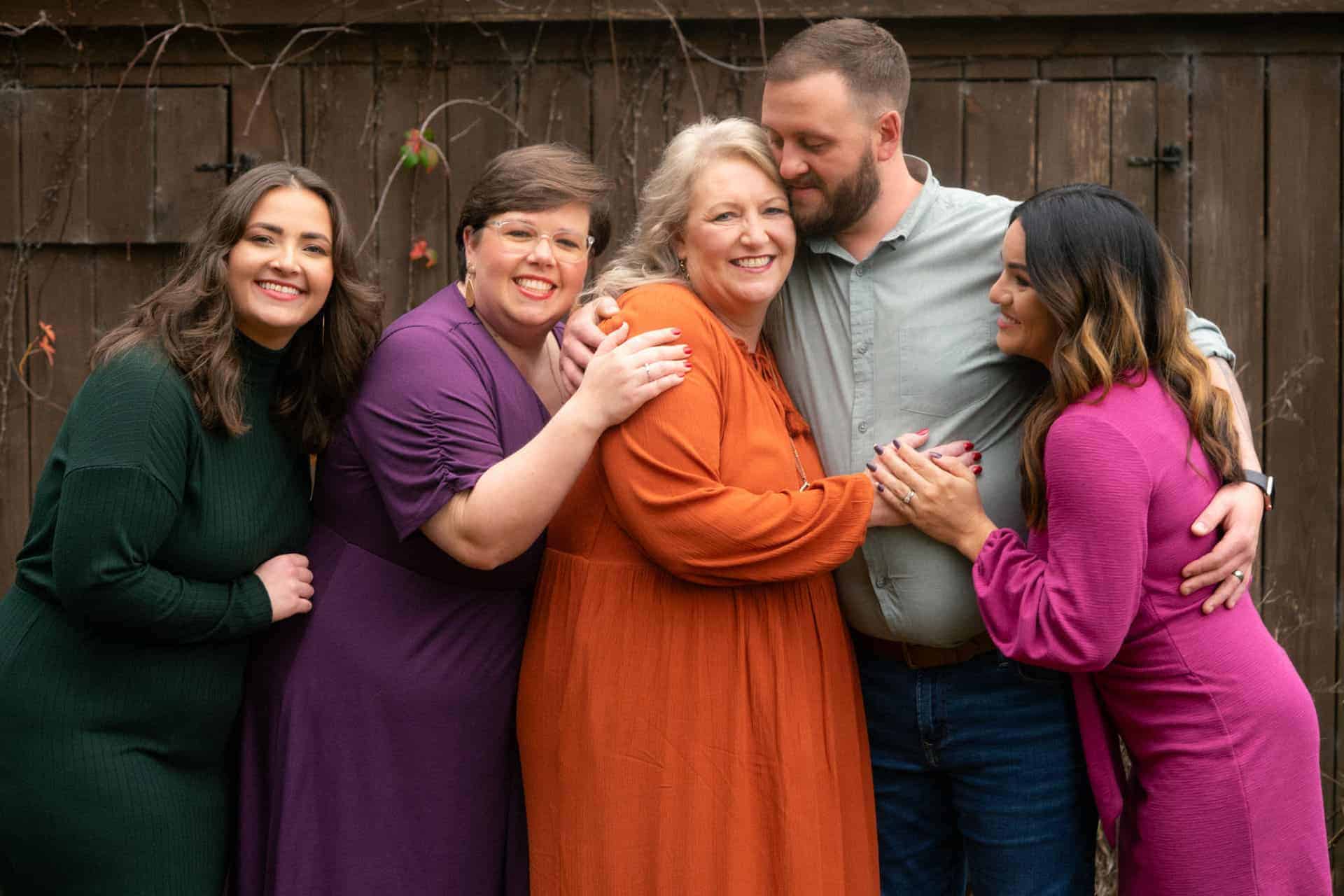 Family portrait with five members smiling and hugging outdoors in front of a wooden fence.