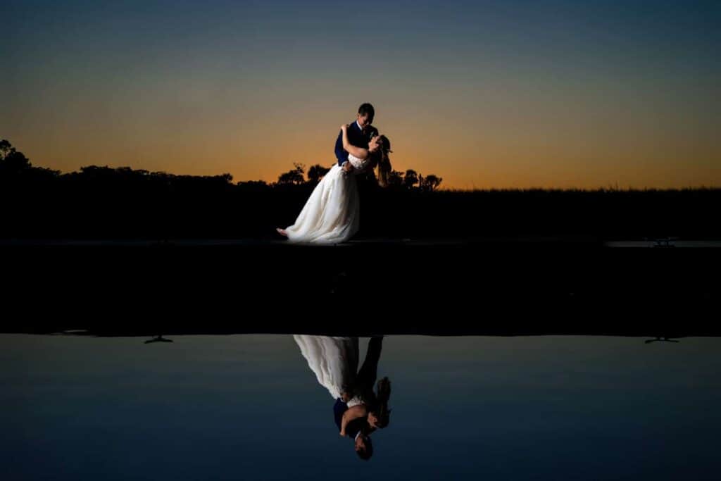 Bride and groom dancing at sunset with reflection in water.