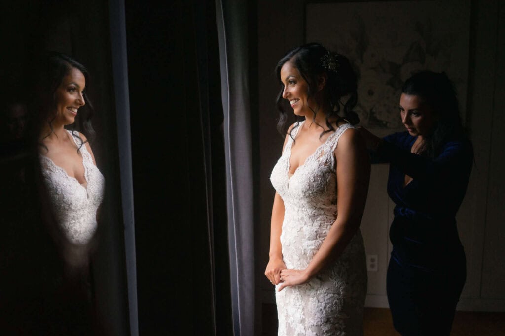 Bride in lace wedding dress smiling at mirror with assistant helping her.
