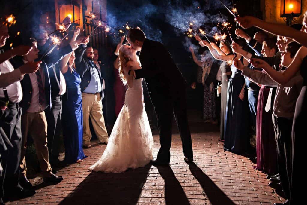 Bride and groom kissing under sparklers during wedding celebration.