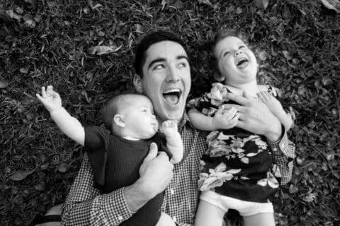 Black and white photo of man holding children while laughing and laying in the grass