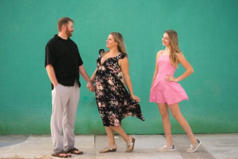 Family including man, woman, and daughter small and look at each other in front of a green wall
