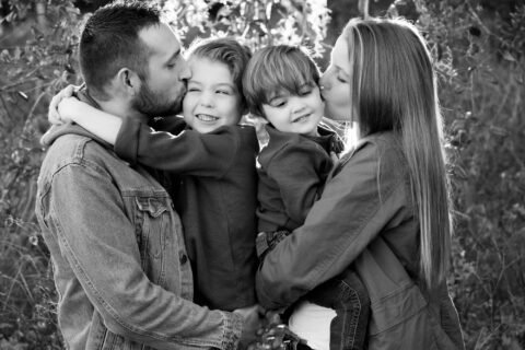 Black and white photo of man and woman hold and kiss their children in front of greenery
