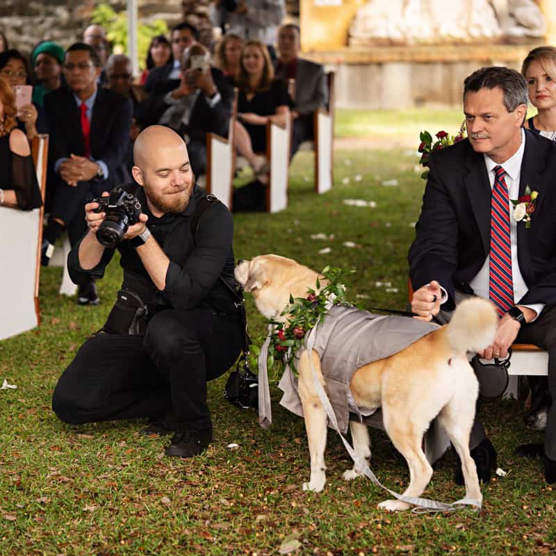 Crouching wedding photographer smiling at a dog during a wedding ceremony