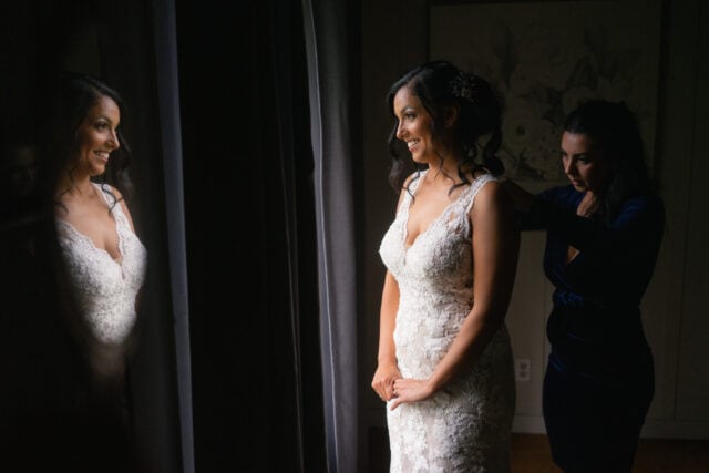 Bridesmaid helps bride with her dress as bride smiles at her relfection
