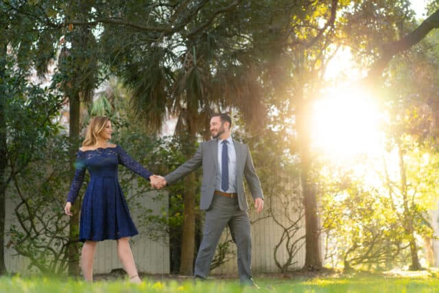 Man and woman hold hands and walk through a park with the sunset in the background