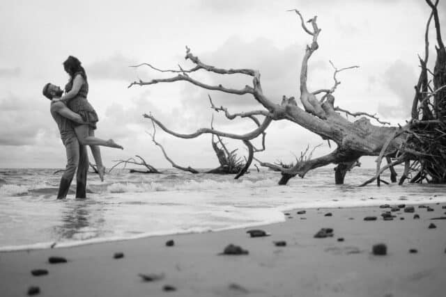 Black and white photo of man holding up woman in the water at the beach with driftwood nearby