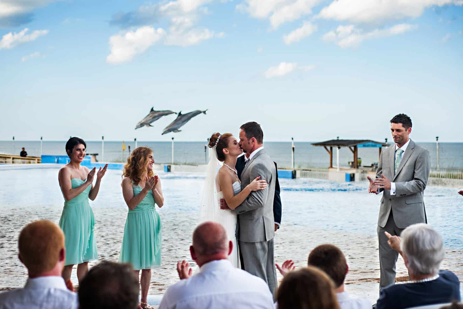 Bride and groom kiss at wedding ceremony while guests clap and dolphins jump in the background