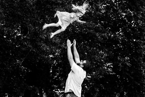 Black and white photo of man throws smiling child into the air in front of greenery